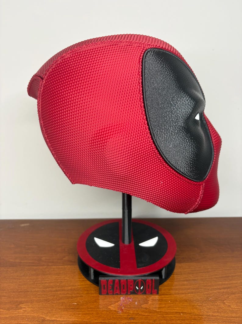 May include: A red and black textured Deadpool helmet displayed on a matching circular stand. The helmet features a prominent black eye patch area. The base of the stand is red and black with white eye accents and a black plaque with the text Deadpool.