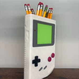 May include: A white pencil holder shaped like a retro handheld gaming device, with a green screen and black and pink buttons. The holder is filled with yellow pencils with pink erasers.