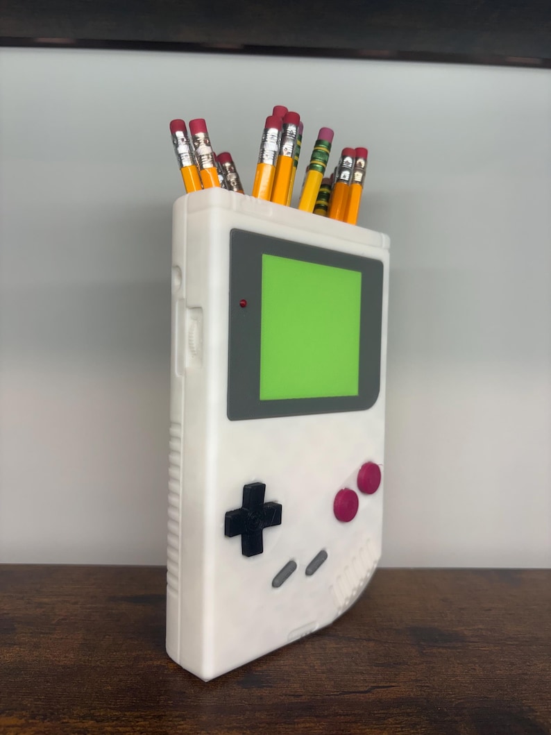 May include: A white pencil holder shaped like a retro handheld gaming device, with a green screen and black and pink buttons. The holder is filled with yellow pencils with pink erasers.