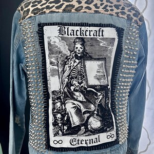 May include: A blue denim jacket with a black and white patch featuring a skeleton figure and the text "Blackcraft Eternal". The jacket has silver studs on the shoulders and back.