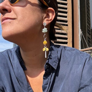 May include: A pair of dangle earrings with a silver chain and multiple beads. The beads are white, yellow, and green. The earrings are hanging from a person's ear.