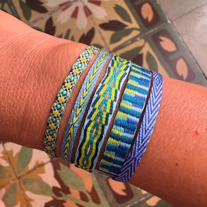 Può includere: Una collezione di braccialetti colorati tessuti in tonalità di blu, giallo e verde. I braccialetti presentano intricati motivi geometrici e sono impilati su un polso. I braccialetti sono in stile bohémien.