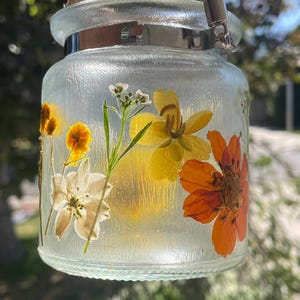 May include: A clear glass jar with pressed flowers in shades of yellow, orange, and white. The jar has a silver metal band and a rope handle. The background is blurred, suggesting an outdoor setting.