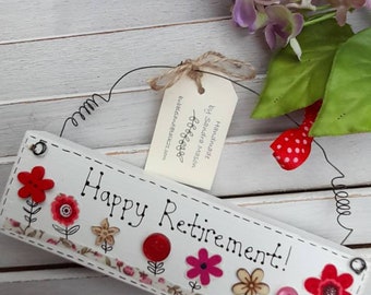 May include: White wooden sign with the text "Happy Retirement!" decorated with red, pink, and cream fabric flowers and buttons.