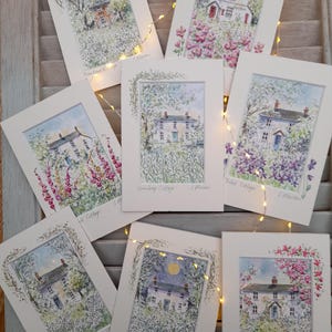 May include: A collection of watercolor art prints featuring charming cottage scenes. Each print, framed in white, depicts a different cottage surrounded by lush gardens and floral elements. The prints are labeled with cottage names like "Snowdrop Cottage" and "Cherry Tree Cottage."