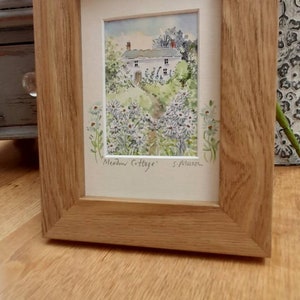Könnte beinhalten: Ein gerahmtes Aquarellbild eines weißen Häuschens mit grünem Dach, umgeben von weißen Blumen und grünem Laub. Das Bild ist mit "Meadow Cottage" betitelt und von "S. Munson" signiert.