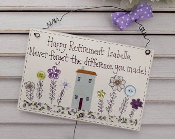 May include: A white wooden sign with a purple bow and the text "Happy Retirement Isabella Never forget the difference you made!" with a small house and flowers. A white wooden flower with the text "Clifton 2021" hangs from the sign.