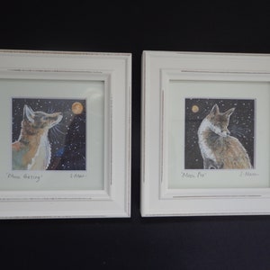 May include: Two framed watercolor paintings of foxes in a snowy landscape. The painting on the left shows a fox looking up at the moon, with the text 'Moon Gazing' and 'S. Mann' below. The painting on the right shows a fox sitting down, with the text 'Mini Fox' and 'S. Mann' below.