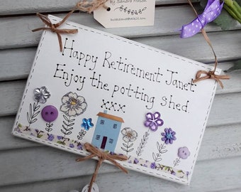 May include: White wooden sign with the text "Happy Retirement Janet: Enjoy the potting shed. XXX" in black cursive writing. The sign is decorated with purple and blue buttons, a small blue house, and a white flower with a blue button in the center.
