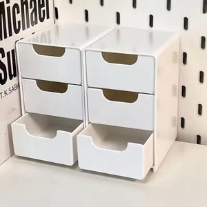May include: Two white plastic desktop organisers, each featuring three stacked drawers with semi-circular cutouts for handles. The units have a smooth, glossy finish and a rectangular frame, with the bottom drawers pulled out to reveal empty interiors.