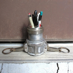May include: A repurposed metal fitting used as a pen holder, filled with various pens and markers. The holder is silver with a textured surface and has two ring handles. The background is a brown door and a concrete step.