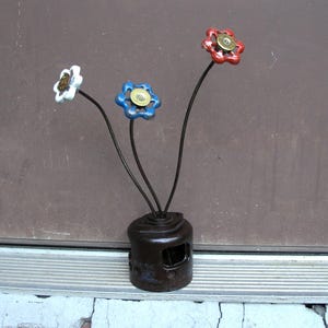 May include: A unique, handcrafted metal sculpture resembling flowers. The sculpture features three flower-like heads in white, blue, and red, each with a brass center, atop dark stems. The base is a repurposed, dark brown metal object. The text "Flowers not included" is at the top.