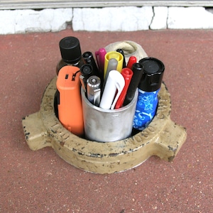 May include: A weathered, beige metal container holding various pens, markers, and a small orange device. The container sits on a red brick surface. Pens are in a silver metal cup. The pens and markers are in various colors, including red, yellow, black, and white.