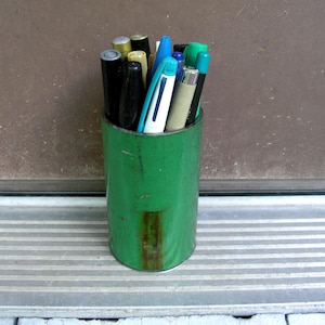 May include: A green cylindrical container filled with various pens and markers. The pens are in a variety of colors, including black, blue, green, and silver. The container is made of metal and shows signs of wear.