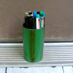 May include: A green cylindrical container holding various pens and markers. The container shows signs of wear with rust-colored streaks. The pens and markers are of different colors, including black, blue, green, and white.