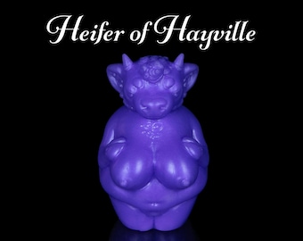 Pode incluir: Uma estatueta roxa de uma criatura semelhante a uma vaca com o texto "Heifer of Hayville" em escrita branca. A estatueta tem um corpo arredondado, pequenos chifres e seios proeminentes, contra um fundo preto.