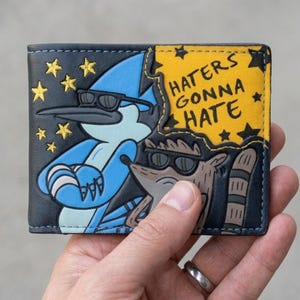 Pode incluir: Uma carteira dobrável com os personagens de desenho animado Mordecai e Rigby de Regular Show, ambos usando óculos de sol. A carteira tem um design azul e amarelo com o texto "Haters Gonna Hate" e estrelas.