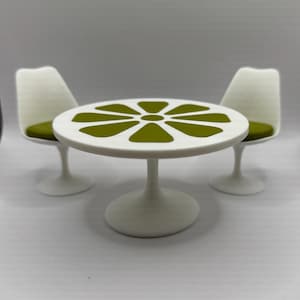 Op de afbeelding: Een miniatuur witte tafel en twee stoelen set. De tafel heeft een rond blad met een groen bloemmotief en een enkele voet. De stoelen hebben witte rugleuningen en groene zitkussens. De set is ontworpen voor een poppenhuis of iets dergelijks.