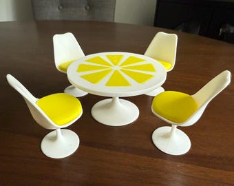 May include: A miniature white table and four chairs set. The table has a white base and a round top with yellow triangular accents. The chairs have white frames and yellow seat cushions. The set is designed to look like a lemon.
