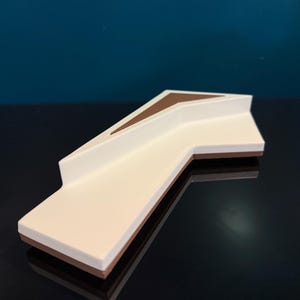 May include: A geometric, L-shaped object featuring a smooth white top surface with a recessed brown triangular accent. The base of the object is brown, creating a layered effect with the white upper section. The design is angular and sharp.
