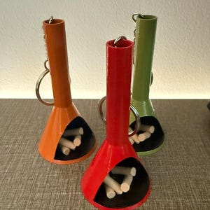 May include: Three colorful miniature fireplace keychains. The keychains are in orange, red, and green, each with a black interior and small wooden logs. The keychains have a silver keyring attached to the top.