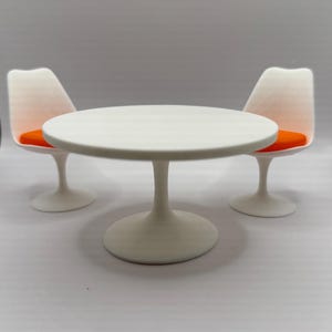 Peut inclure: Une table ronde blanche avec un pied central, flanquée de deux chaises blanches. Les chaises ont un dossier incurvé et des coussins de siège orange. L'ensemble de meubles est de style moderne.