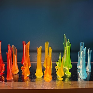 Op de afbeelding: Een verzameling kleurrijke glazen vazen in een regenboog van tinten, waaronder rood, oranje, geel, groen en blauw. De vazen variëren in vorm en grootte, met sommige met een slank, langwerpig ontwerp en andere met een rondere vorm. De vazen zijn gerangschikt op een reflecterend oppervlak.