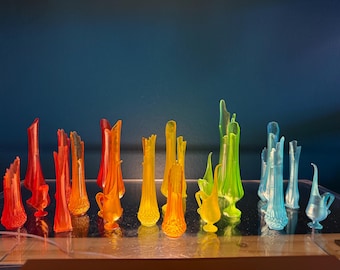 May include: A selection of vibrant glass vases in a spectrum of colours, including red, orange, yellow, green, and blue. The vases have diverse shapes and sizes, with some being slender and elongated, while others are more rounded. The vases are displayed on a reflective surface.