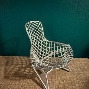 May include: A miniature silver-colored chair featuring a wire grid seat and backrest design. The chair is constructed with a metallic frame and angled legs, showcasing a distinct lattice pattern throughout the seating area and back support structure.