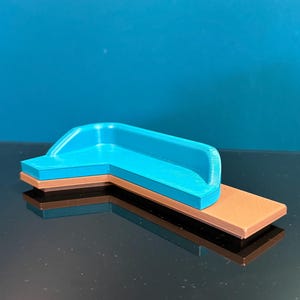 May include: A miniature L-shaped sofa model featuring a bright cyan blue seat and backrest section mounted on a flat, rectangular tan base. The object has a smooth, layered texture consistent with 3D printed plastic construction and a clean finish.