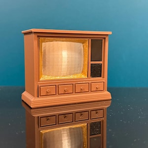 May include: A miniature television set in a light brown plastic finish. The unit features a gold-colored screen area, two black speaker panels on the right side, and four small drawers with circular knobs along the bottom front section.