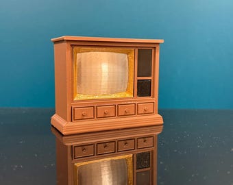 May include: A miniature television set in a light brown plastic finish. The unit features a gold-coloured screen area, two black speaker panels on the right side, and four small drawers with circular knobs along the bottom front section.
