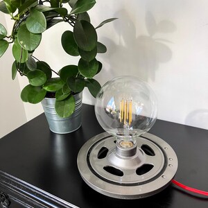 Peut inclure: Une lampe de table en métal gris avec un globe en verre transparent. La lampe a une base ronde avec un motif circulaire et un cordon rouge.