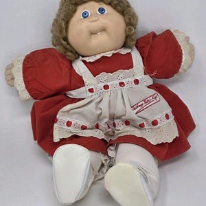 May include: A vintage Cabbage Patch Kids doll with curly brown hair and blue eyes. The doll wears a red dress with a white apron featuring a red berry design and the text "Cabbage Patch Kids". White shoes and socks complete the outfit.