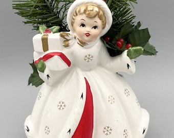 May include: A ceramic figurine of a person wearing a white hooded coat with red trim and gold snowflake patterns. The figure holds a white gift box with a red ribbon. Behind the figurine are green pine needles and small red holly berries.