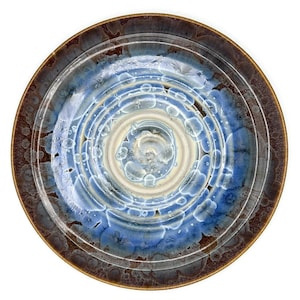 May include: A round ceramic plate featuring a crystalline glaze with concentric rings. The center is a creamy white, transitioning into vibrant blue and white crystalline patterns, surrounded by a dark brown rim with textured crystalline details.