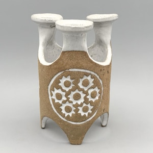 May include: A ceramic triple-spout vase featuring a textured tan exterior with a circular white relief pattern of seven star-like shapes. The upper rims and interior of the three openings are finished in a smooth white glaze, contrasting with the tan body.
