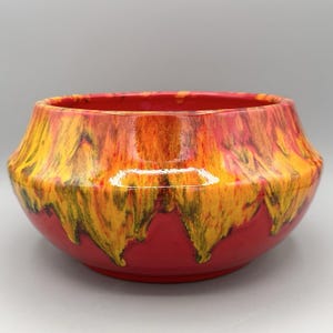 May include: A ceramic bowl featuring a vibrant glaze with a deep red base color and a flowing, drip-like pattern of bright yellow and orange hues cascading from the rim. The bowl has a wide, rounded body with a smooth, glossy finish throughout.