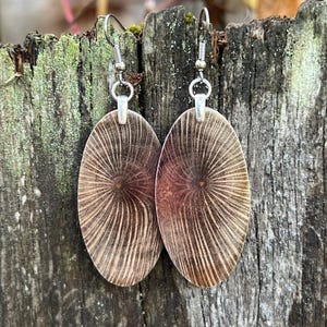 May include: A pair of oval-shaped earrings with a radial pattern in shades of brown and tan. The earrings are suspended from silver-colored hooks. The background is a weathered wooden surface.