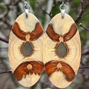 May include: A pair of oval-shaped wooden earrings with a light brown and dark brown wood grain pattern. The earrings have a silver hook and a clear resin center.