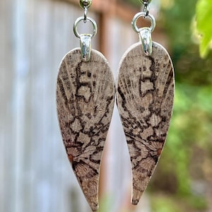 May include: A pair of earrings with a leaf-like shape, featuring a detailed pattern in shades of brown and black. The earrings are suspended from silver-colored hooks. The background is blurred, with green foliage and a wooden branch.