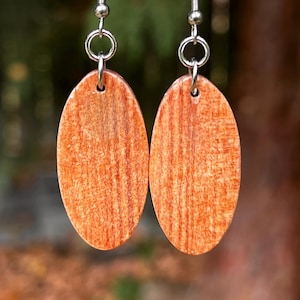 May include: A pair of oval-shaped earrings featuring a natural orange-toned wood grain pattern. Each earring is suspended from a silver-toned metal hook by a small circular jump ring, showcasing a smooth, flat surface with visible vertical wood texture.