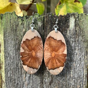 May include: Oval wooden earrings with a natural wood grain pattern. The earrings feature a light-colored wood border with a darker, radial design in the center. Silver-colored ear wires complete the design. The earrings are displayed against a weathered wooden background.