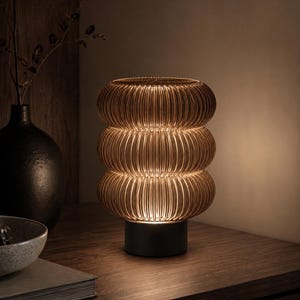 May include: A table lamp featuring a stack of three rounded, ribbed amber glass segments that emit a warm glow. The glass structure is supported by a solid, cylindrical black base, creating a distinct layered appearance with vertical lines.