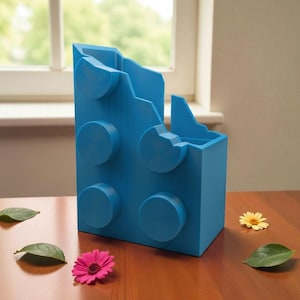 May include: A blue storage container in the shape of a large building block. It features four circular studs and a notched top. A pink flower, a yellow flower, and green leaves are on the table, adding a touch of colour.