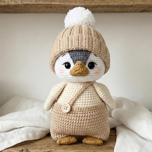 May include: A handmade crochet penguin doll wearing a tan beanie with a white pom-pom, overalls, and matching booties. The penguin has a white face, grey head, and a yellow beak. The doll is approximately 20 cm tall.