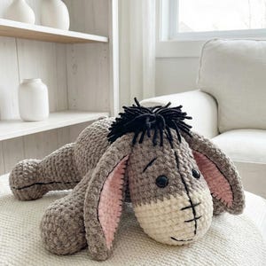 May include: A handmade crochet plush toy of a donkey, featuring a gray body, cream-colored muzzle, and pink inner ears. The donkey has black eyes and a black mane. It is lying on a white surface.