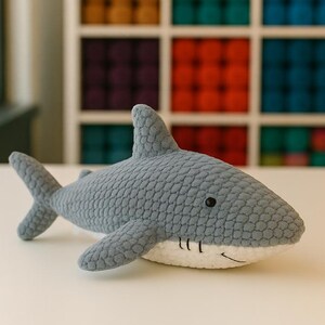 May include: A gray crocheted shark plush toy with a white belly and black eyes. The shark has fins and a tail, and is lying on a white surface. The background features a colorful display of yarn.