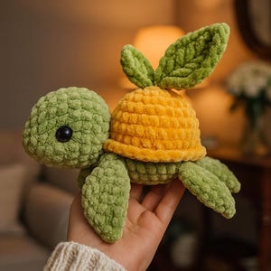 Puede incluir: Un peluche de tortuga hecho a mano con ganchillo en tonos verdes y amarillos. La tortuga tiene un caparazón, cabeza y aletas texturizadas, con un caparazón amarillo brillante y dos acentos verdes en forma de hoja en la parte superior. El ojo de la tortuga es una pequeña cuenta negra.