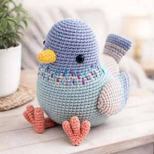 Puede incluir: Un pájaro de ganchillo hecho a mano en tonos de azul, turquesa y beige. El pájaro tiene un pico amarillo, ojos negros y patas de color coral. El pecho presenta un patrón de puntadas decorativas con detalles morados y turquesas. Un juguete suave y tierno.